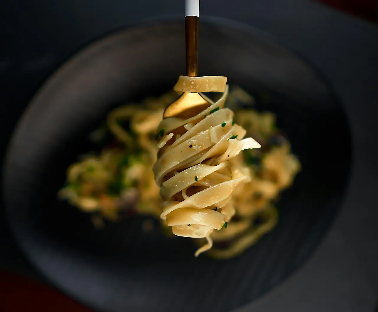 Ribbons of fresh pasta twirled on a fork above a dark ceramic plate, with parmesan shavings and chopped herbs