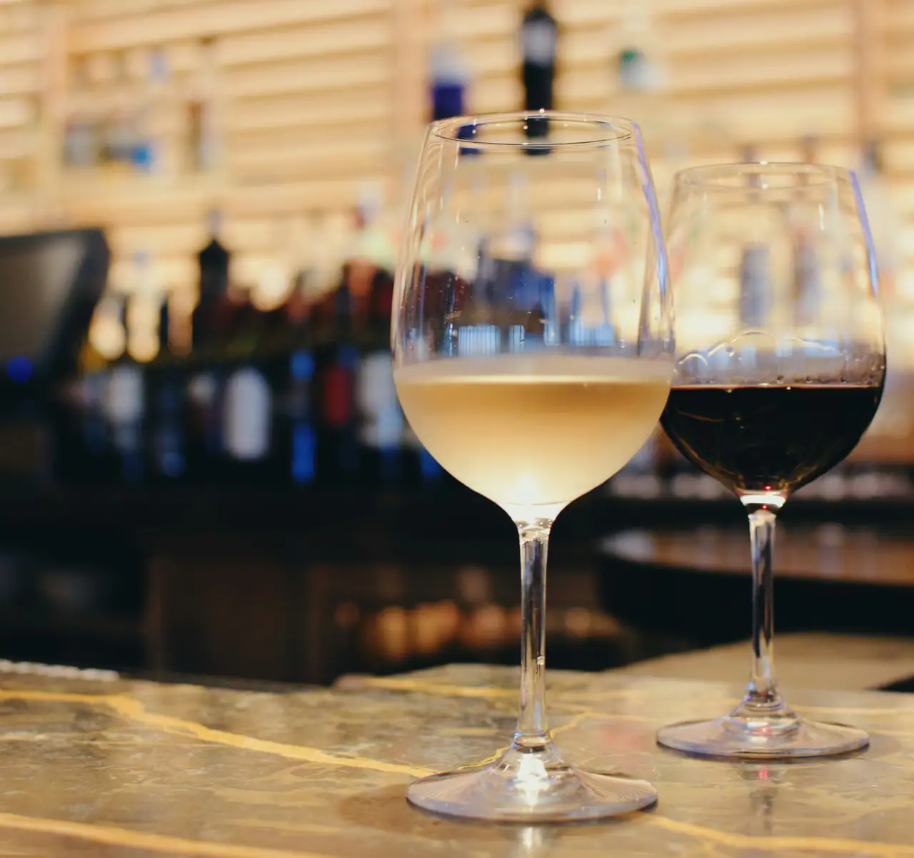 A glass of white wine and a glass of red wine on a stone bar top, with bottles blurred in warm light behind
