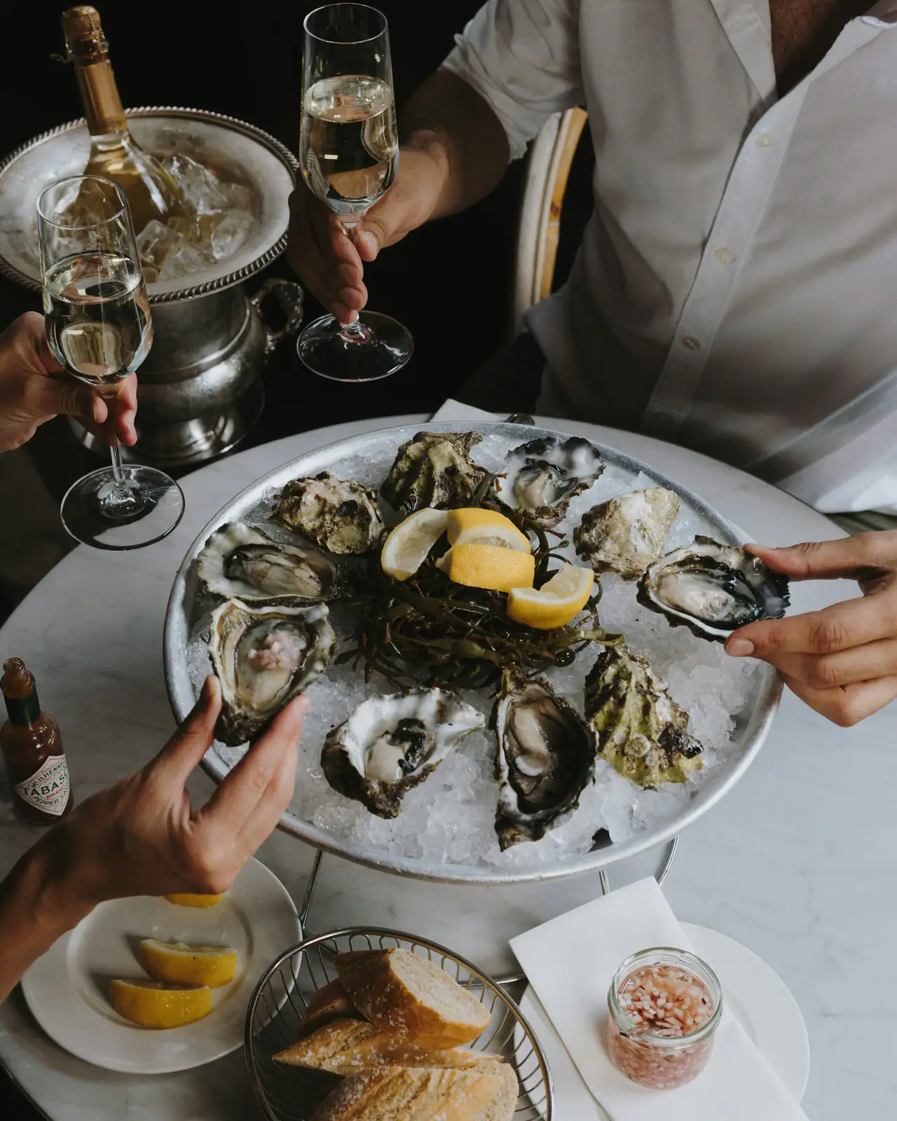 A silver platter of freshly shucked oysters on ice with lemon wedges on a marble table, two glasses of sparkling wine and a champagne bucket beside it