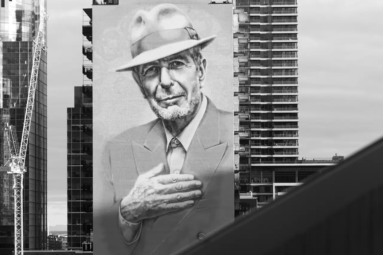 Black-and-white photo of the giant Leonard Cohen portrait mural on a Montréal high-rise, hand on chest, beneath an overcast sky