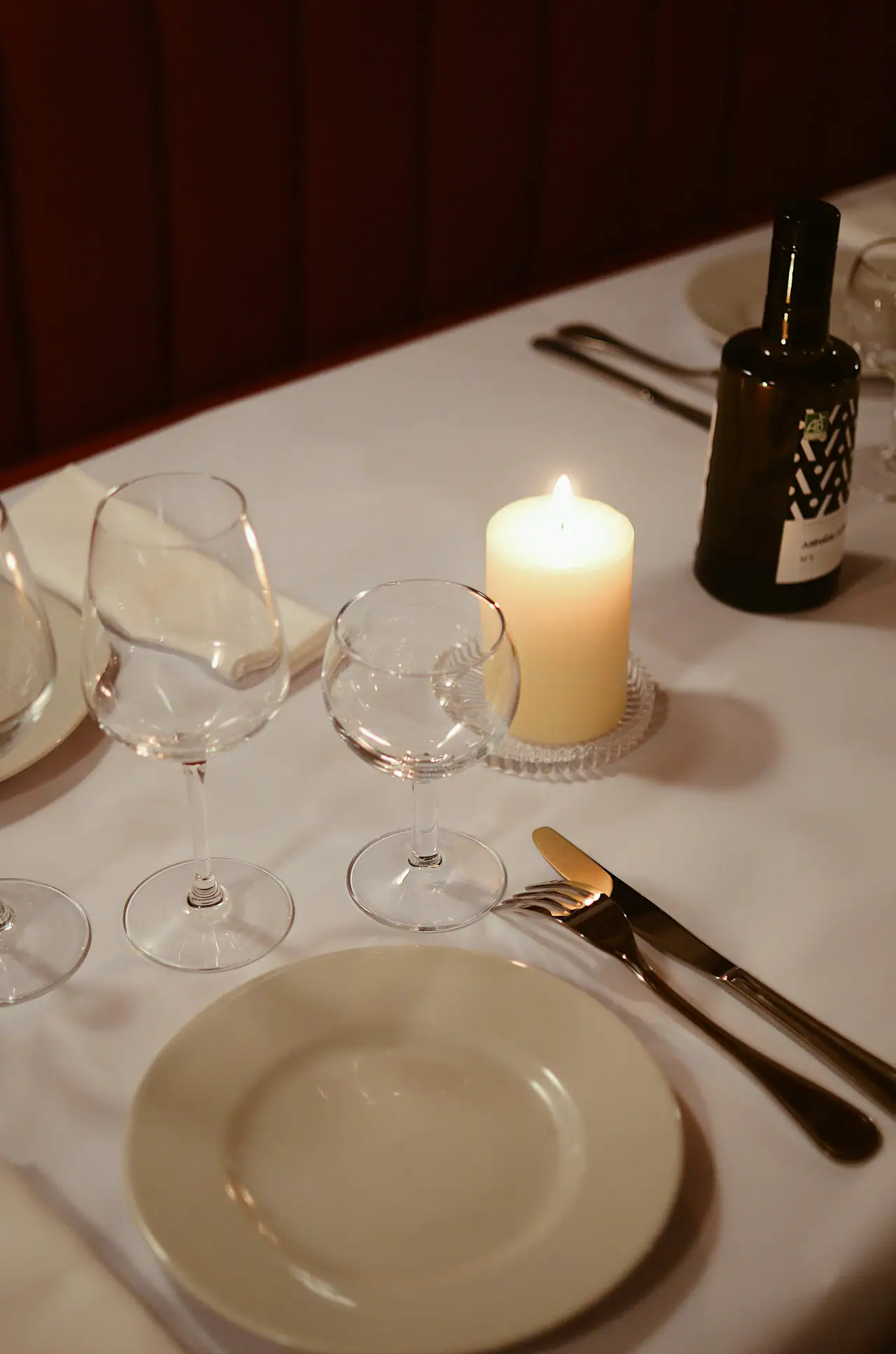 A lit ivory candle between two empty wine glasses on a white tablecloth, with a place setting, an olive oil bottle and a dark red banquette behind