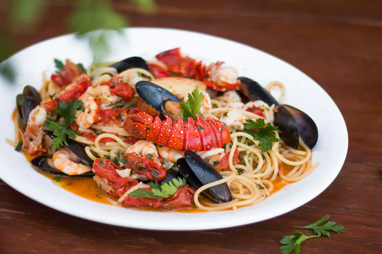 A white plate of spaghetti with lobster tail, mussels, prawns and tomato on a dark wooden table