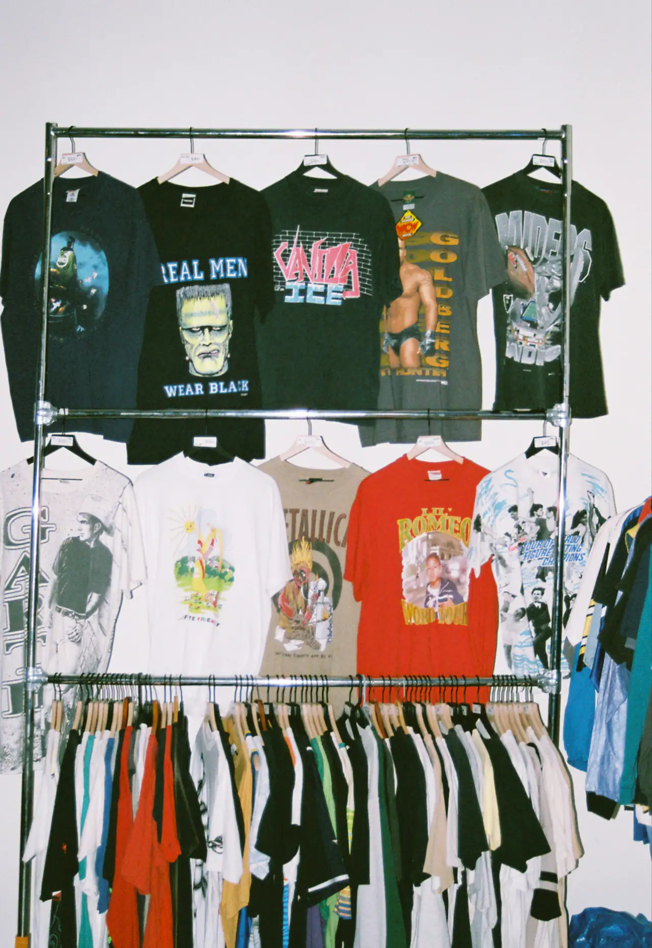 A steel rail of vintage graphic band and wrestling tees on hangers against a white wall, with more shirts crowded on the rail below