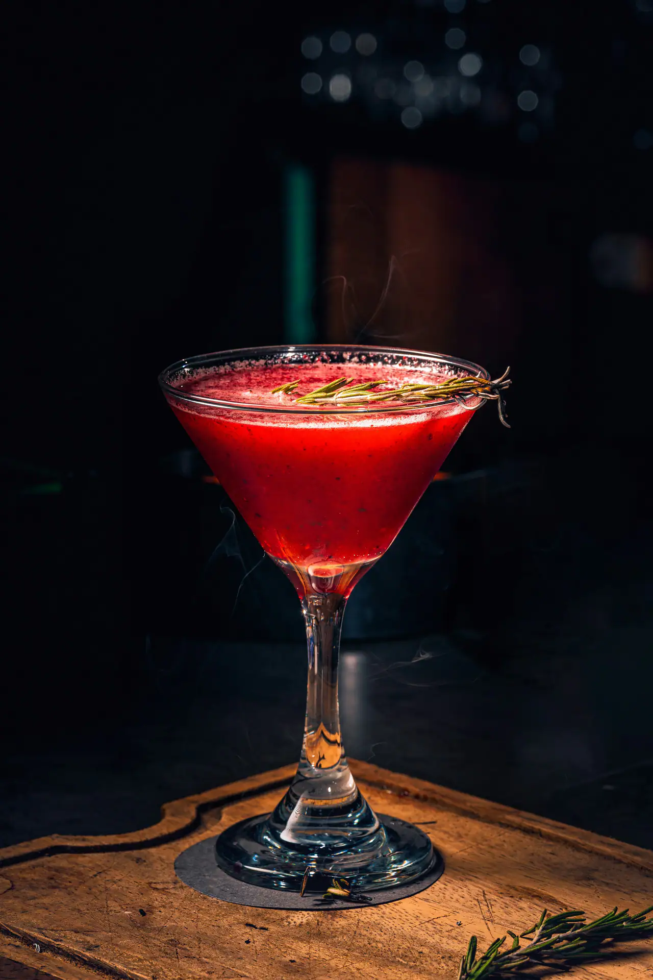 A deep red cocktail in a coupe glass garnished with a sprig of rosemary, wisps of smoke rising, on a wooden board in a dark bar