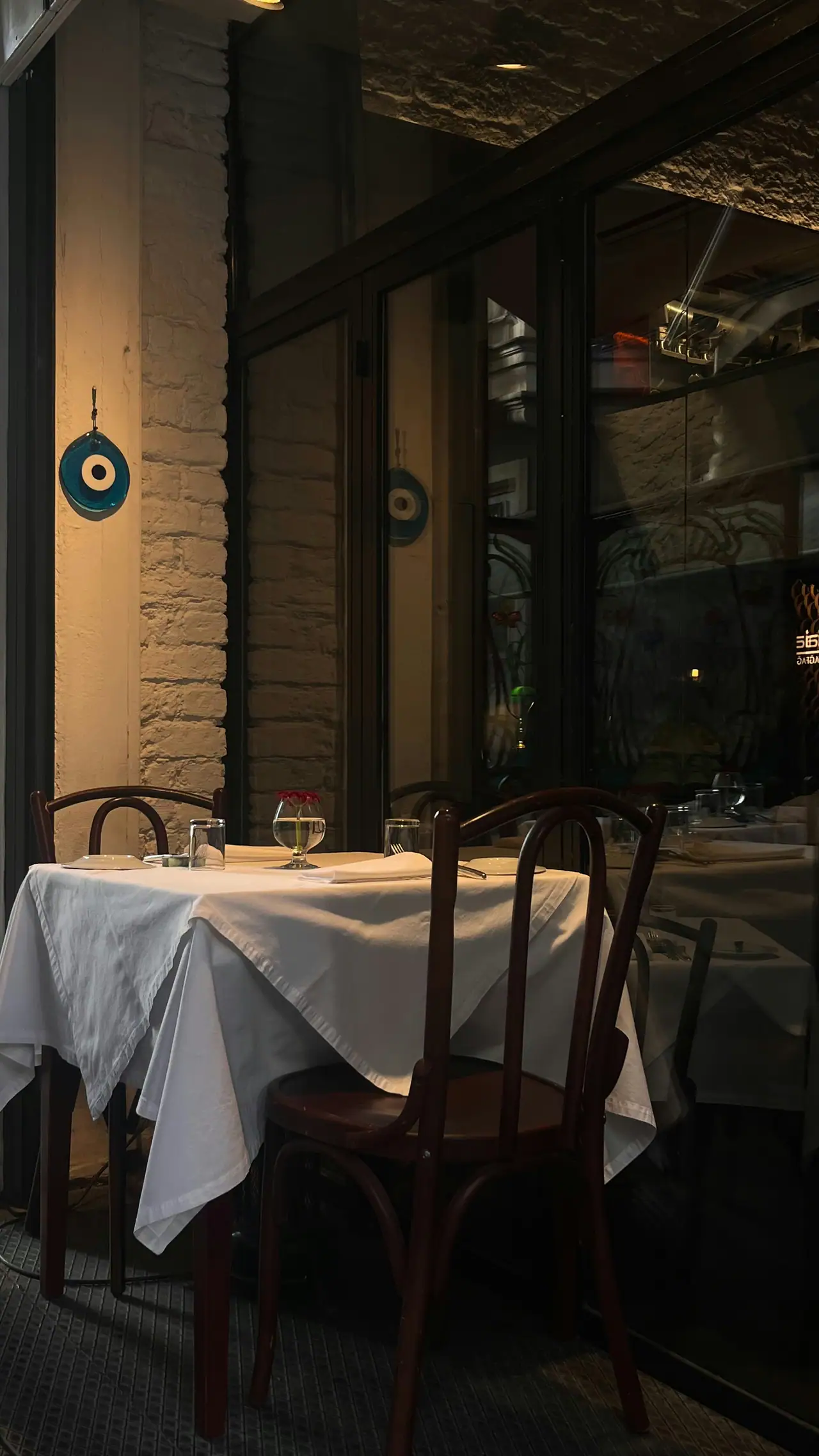A candlelit-looking corner table with white linen, bentwood chairs and a small glass vase, a blue evil-eye charm glowing on a brick wall beside a dark glass window