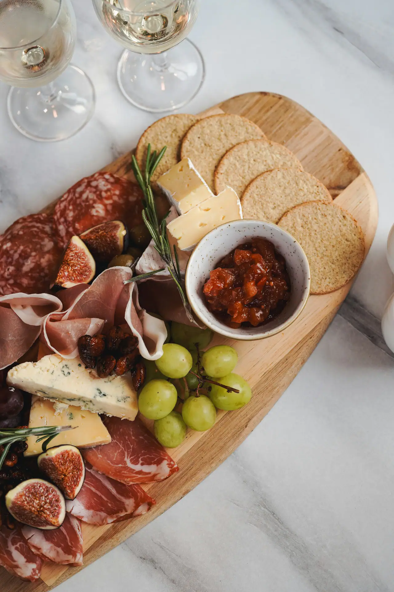 Overhead view of a wooden board with cured meats, figs, soft and blue cheeses, crackers, grapes and chutney beside two glasses of white wine on a marble table