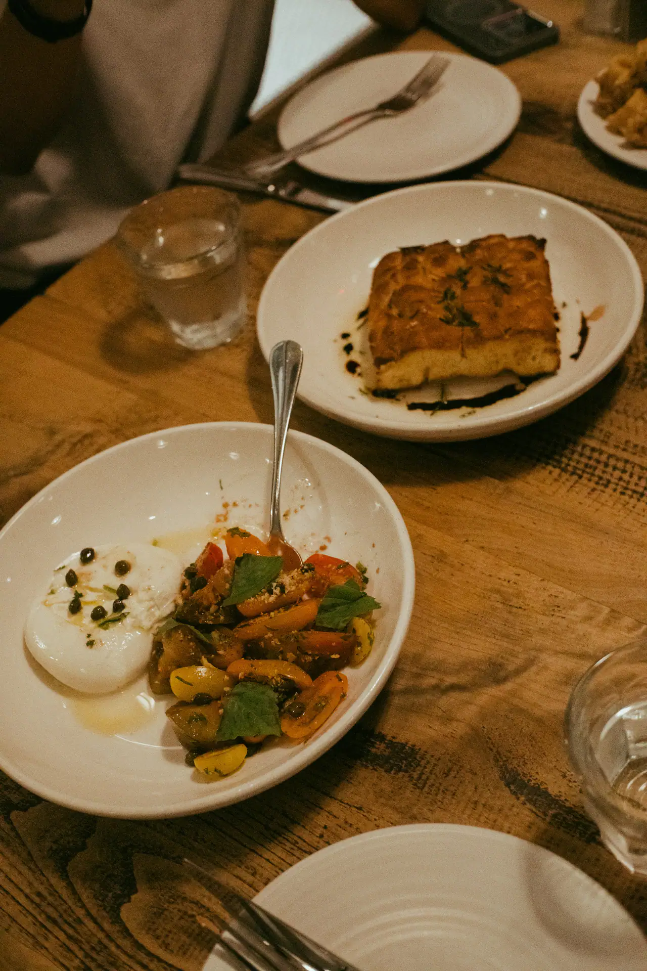 A bowl of burrata with blistered cherry tomatoes, basil and capers beside a plate of focaccia on a worn wooden restaurant table
