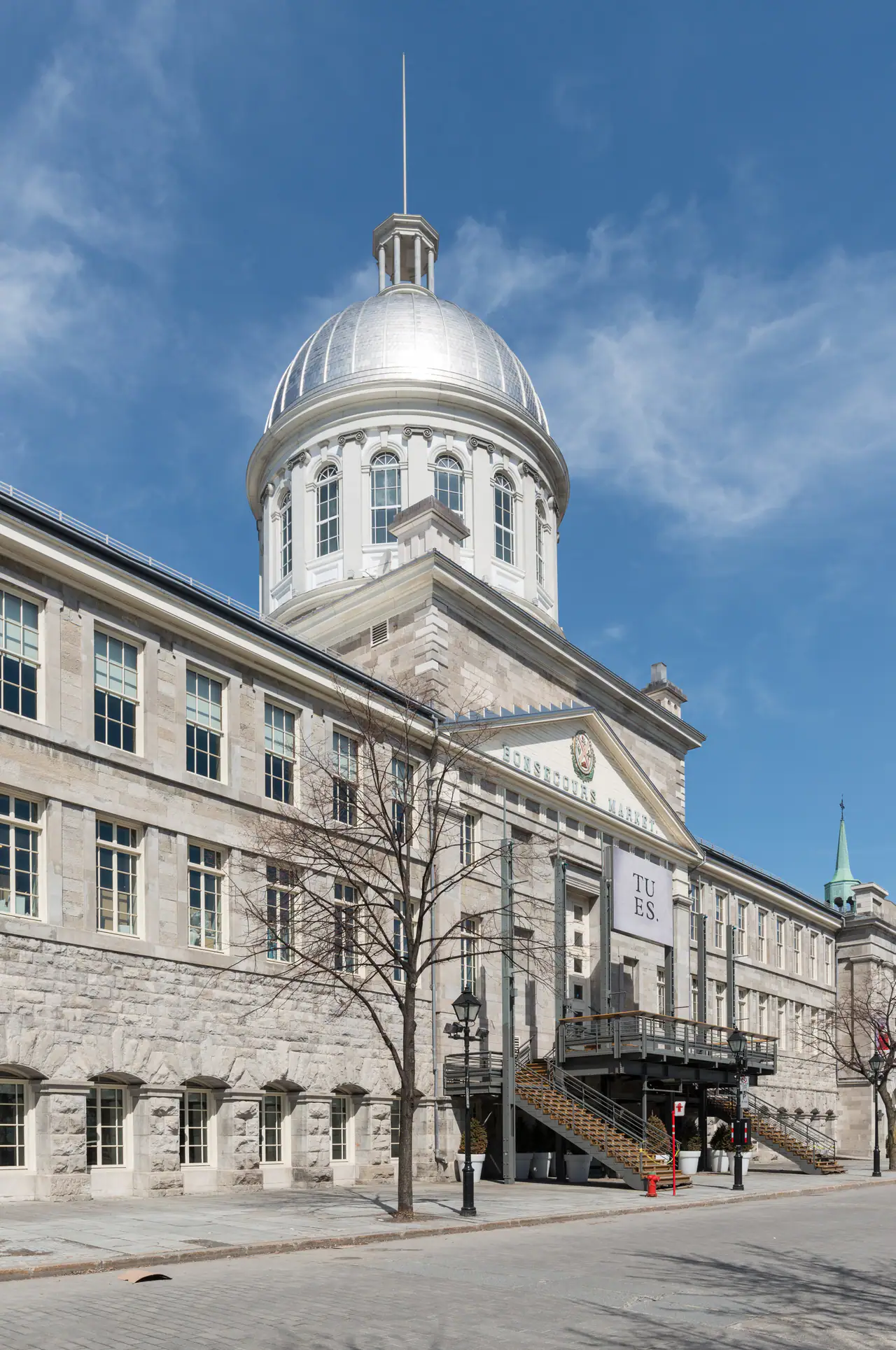The silver dome and grey limestone façade of Marché Bonsecours in Old Montréal under a blue sky, with a bare tree in front