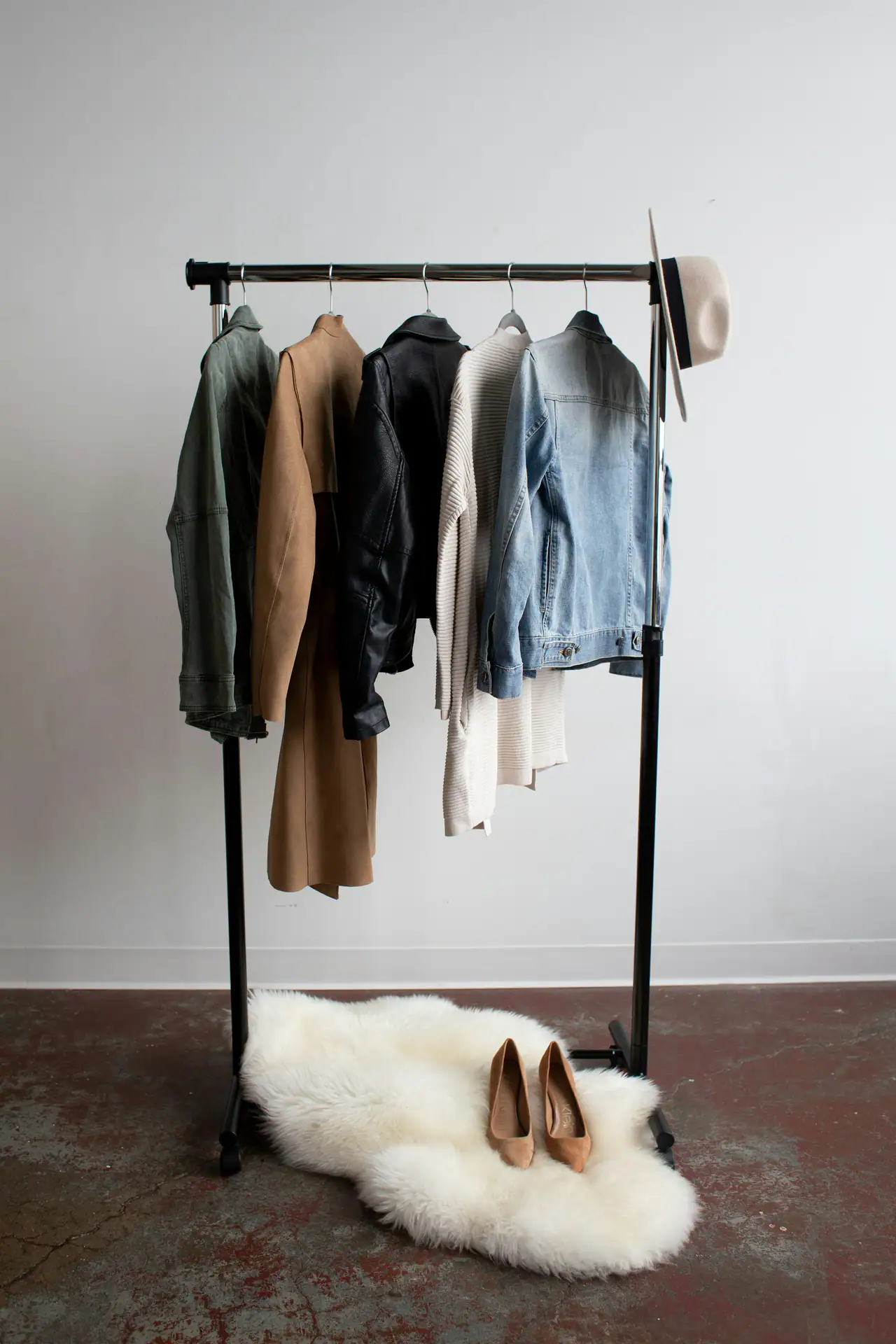 A single clothing rack holding a green jacket, camel suede coat, black leather jacket, cream knit and a light denim jacket, with a felt hat, tan heels and a sheepskin rug on a concrete floor