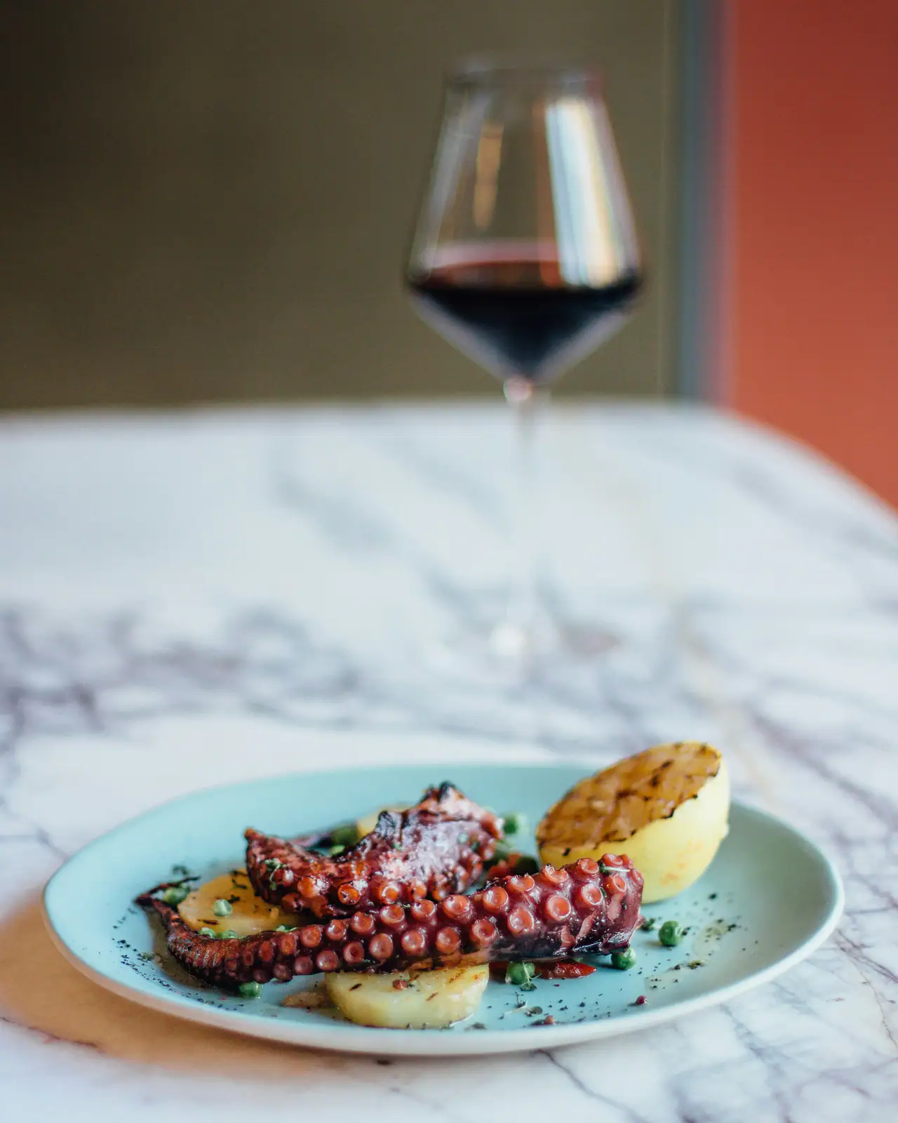 Grilled octopus tentacles with potato and peas on a pale blue plate, on a marble table with a glass of red wine behind