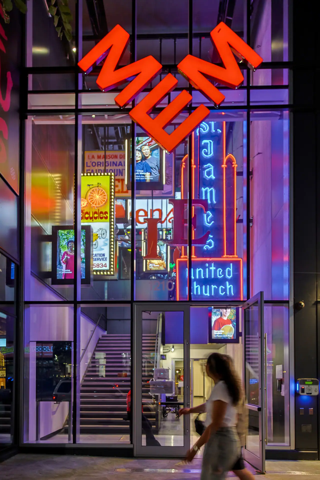 Glass entrance of the MEM museum on Boulevard Saint-Laurent at dusk, with the orange MEM sign above vintage neon signs, including St. James United Church, glowing inside and a blurred passer-by