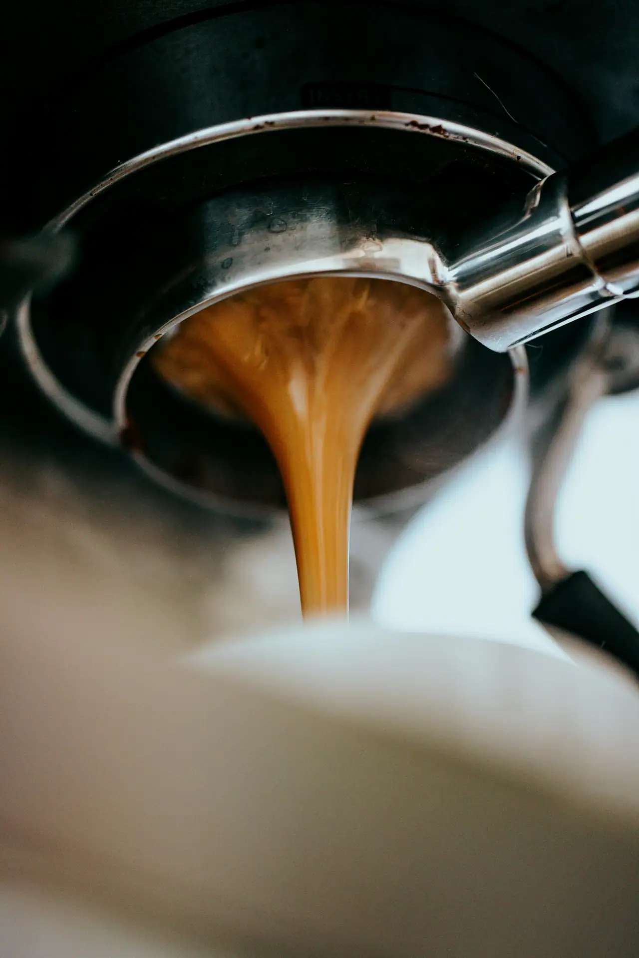 Close-up of espresso pouring from a metal portafilter into a white cup, with dark steel and warm amber tones