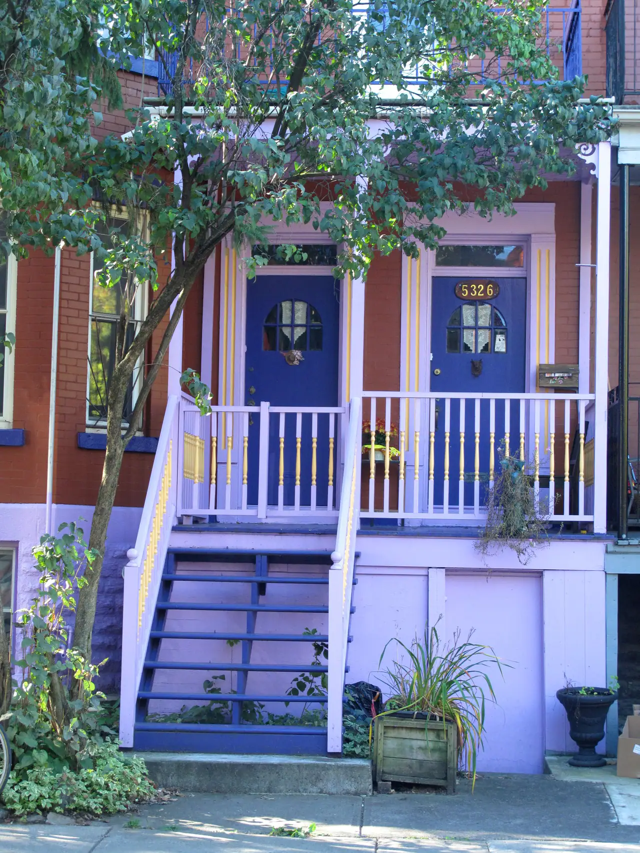 A Mile End duplex with brick facade, lilac-painted porch and stairs, and twin blue front doors beneath a leafy tree