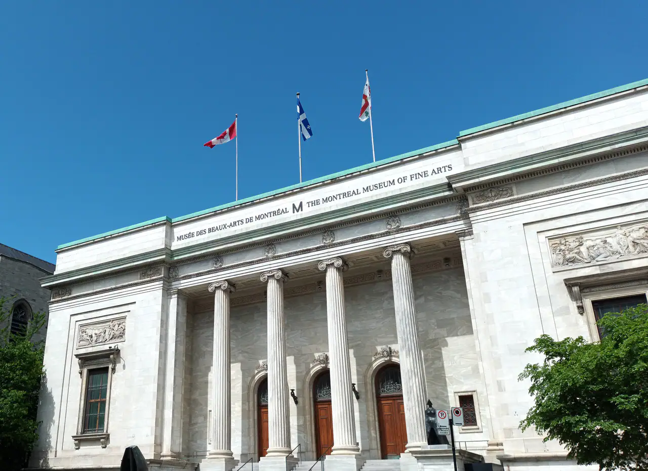 The white marble Beaux-Arts facade of the Montreal Museum of Fine Arts on Sherbrooke Street, with four Ionic columns, arched wooden doors and three flags against a clear blue sky