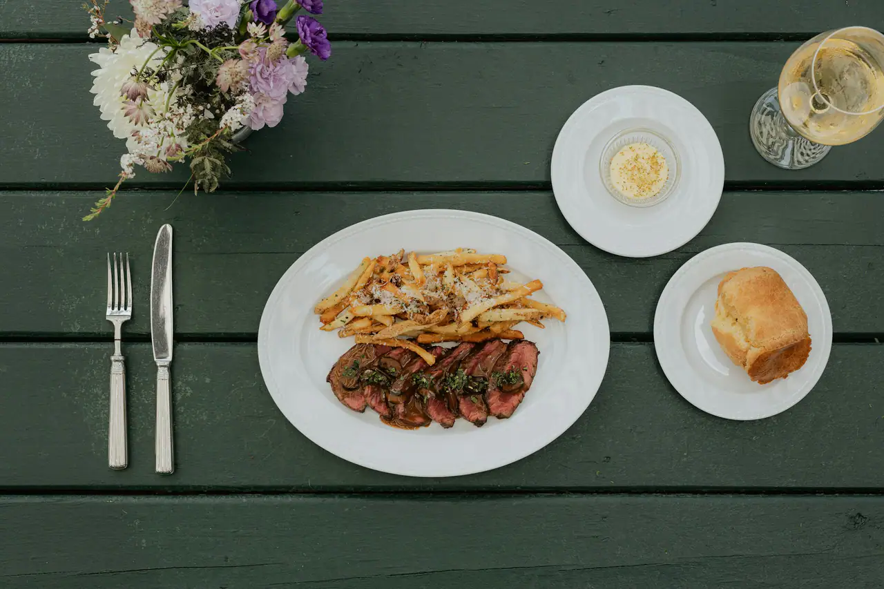 Overhead view of sliced steak with french fries on an oval white plate, beside a glass of white wine, a bread roll and a small bouquet, on a green wooden table