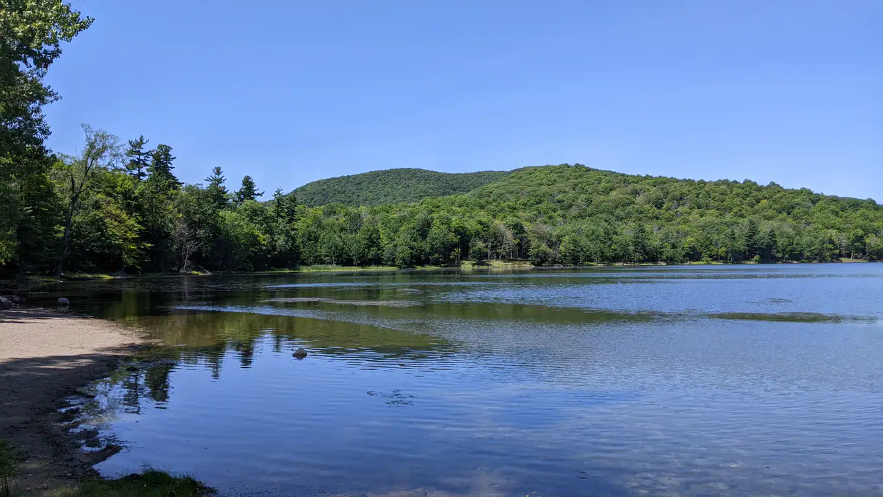 Lac Hertel in the Gault Nature Reserve, with a sandy shore in the foreground and forested summits of Mont Saint-Hilaire rising across the still water under a clear sky