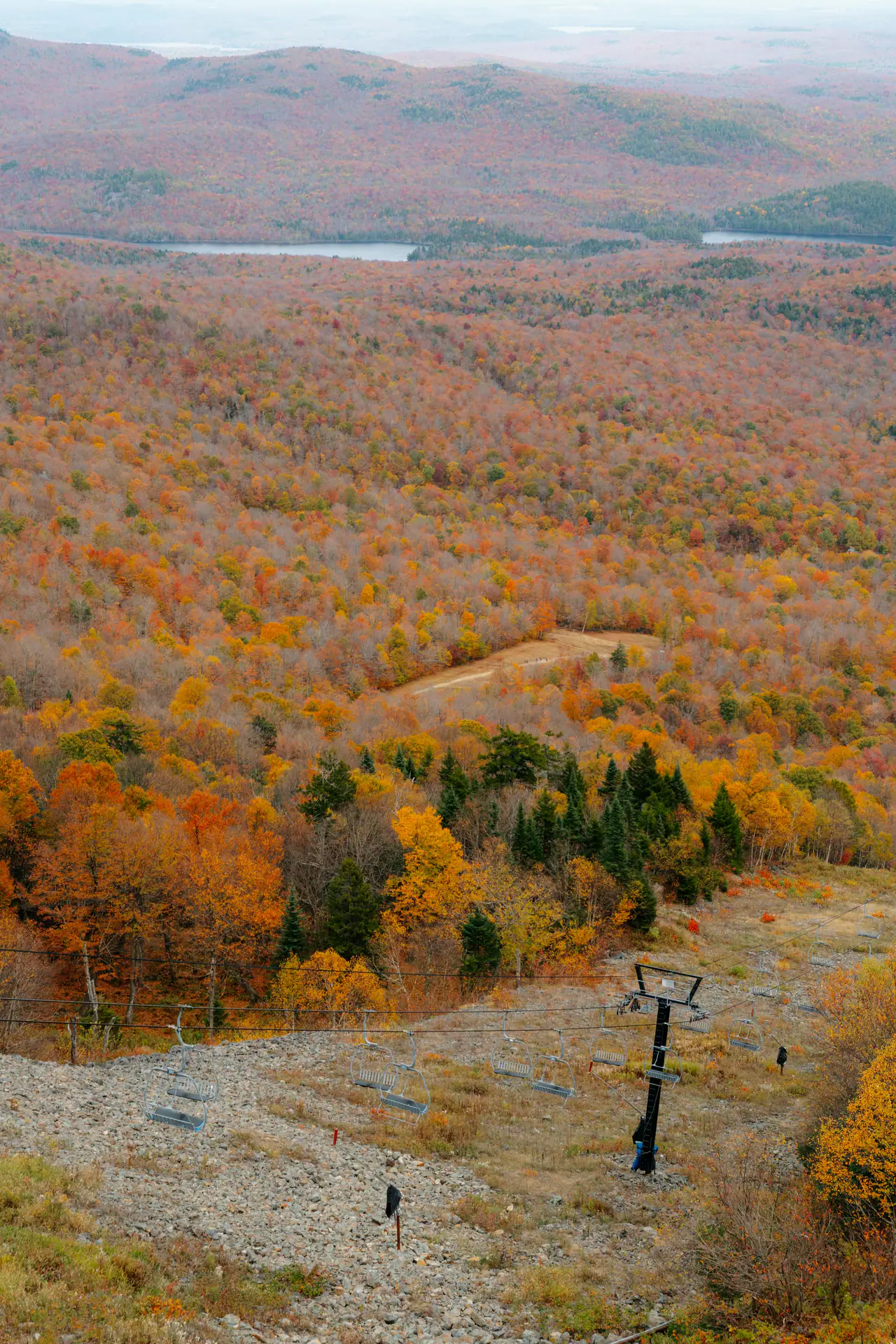 Looking down a ski hill in autumn, an idle chairlift line crossing rocky ground above a wide valley of orange, gold and rust foliage with small lakes in the distance.