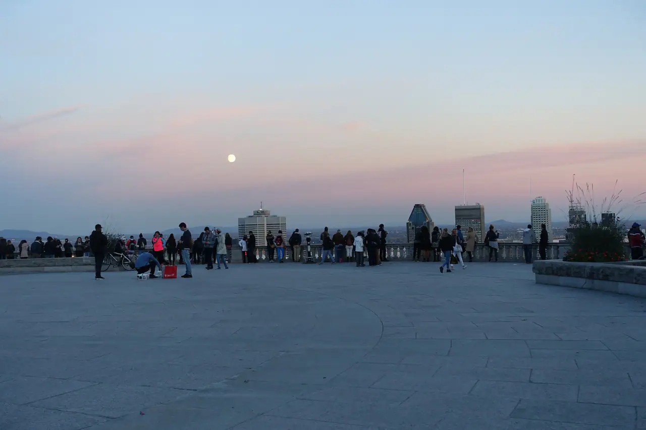 Visitors gathered along the stone balustrade of the Kondiaronk Belvedere on Mount Royal at dusk, with the downtown towers and a pale moon under a pink and lavender sky