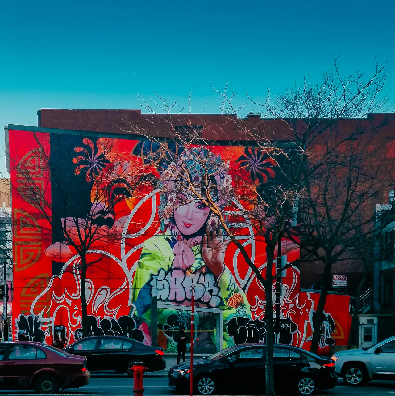 A large street-art mural of a figure in an ornate headdress in red, green and white on a brick building's side wall, seen across a street through bare trees