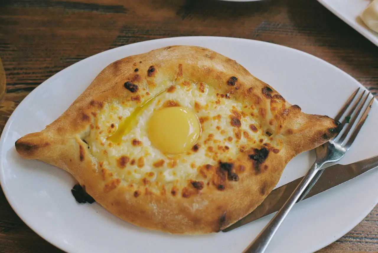 A khachapuri cheese boat with a runny egg yolk on a white oval plate, with a fork and knife, on a dark wooden table