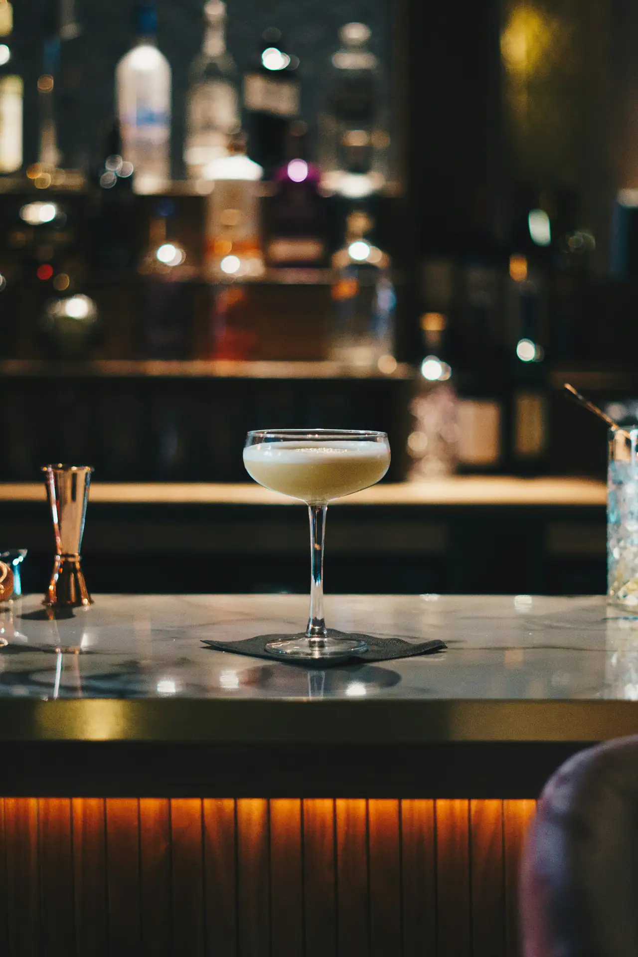 A frothy cream-coloured cocktail in a coupe glass on a marble-topped bar, with a copper jigger and softly blurred bottles in a dim, warm-lit room behind