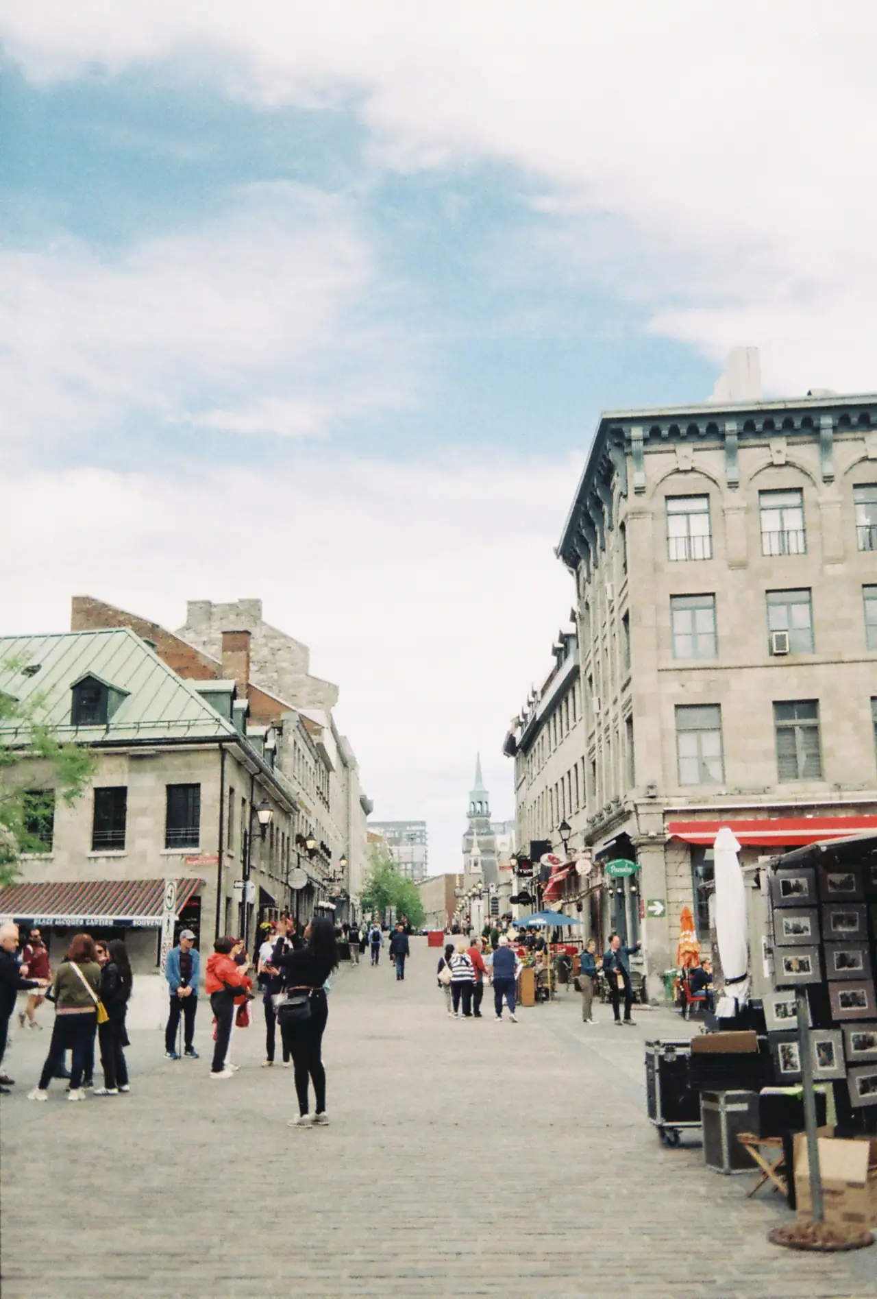A cobblestone street in Old Montréal lined with grey stone buildings, with a church spire in the distance under a soft cloudy sky