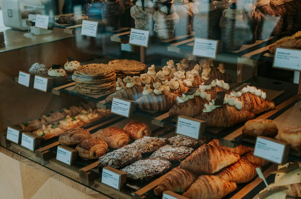 A wood-framed glass pastry case holding croissants, almond croissants, cookies and cream-topped pastries with small price cards
