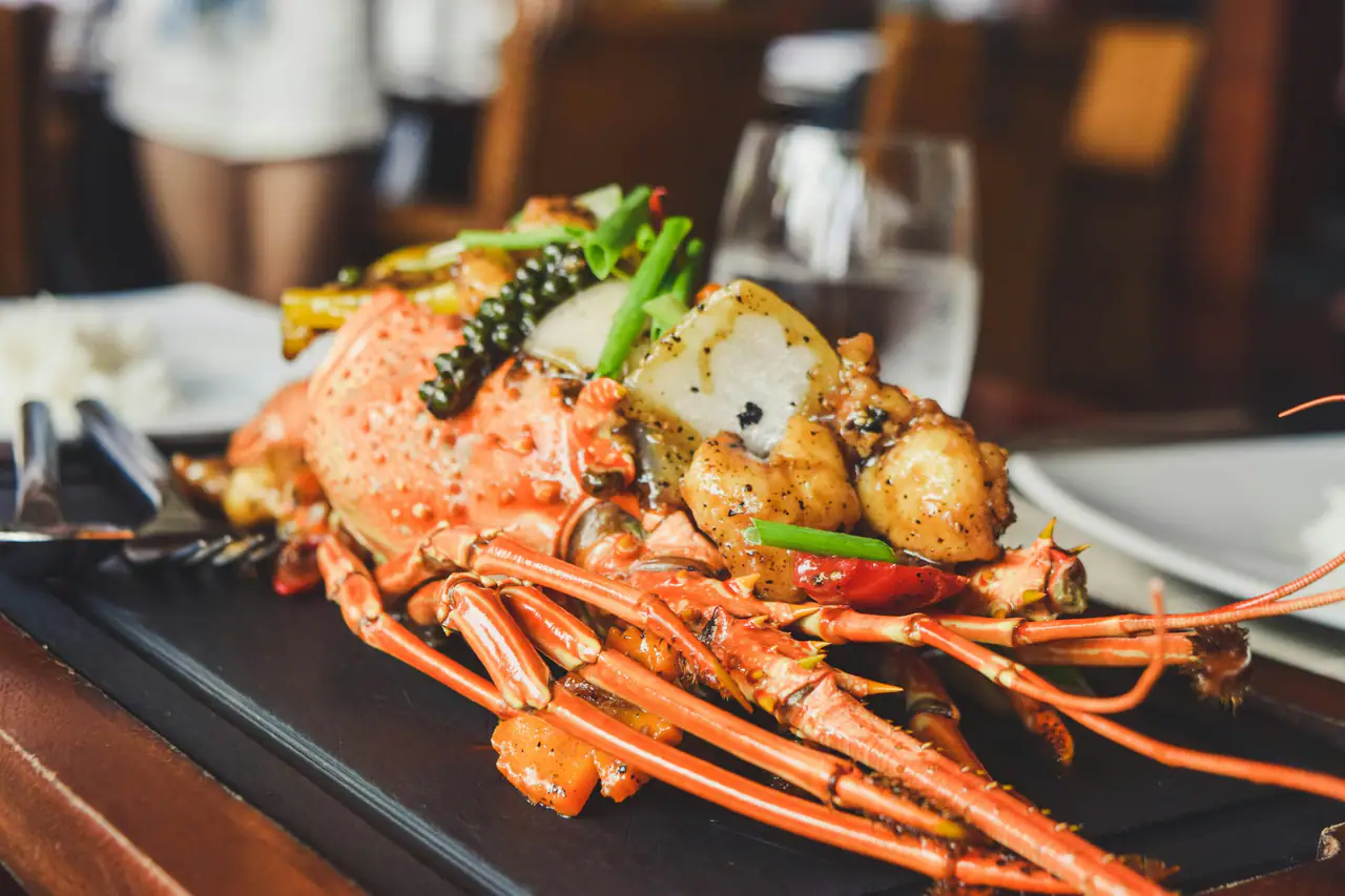 A whole stir-fried lobster with green onion, chili and garlic served on a dark slate tray in a warmly lit restaurant, a water glass blurred behind
