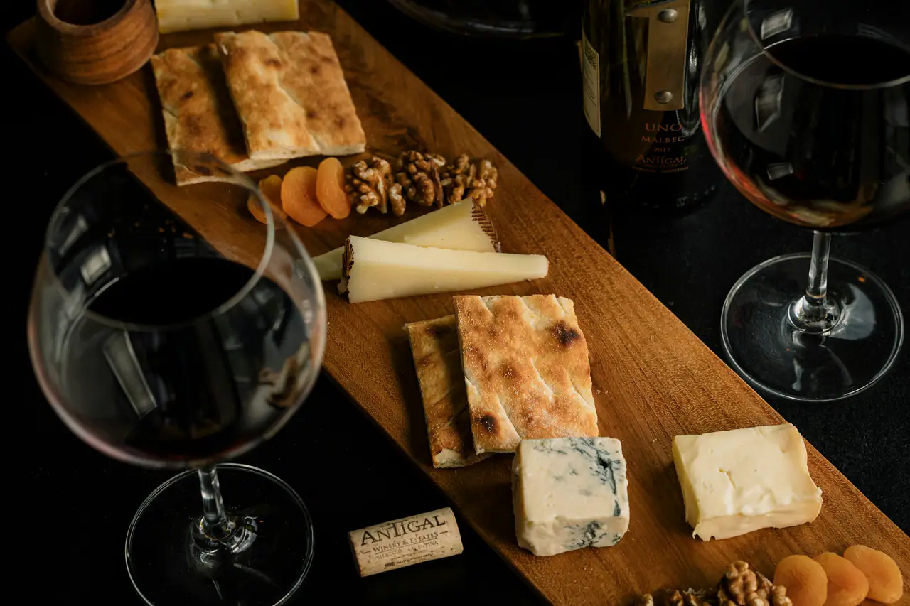Two glasses of red wine beside a wooden board of crackers, aged cheeses, walnuts and dried apricots on a dark table