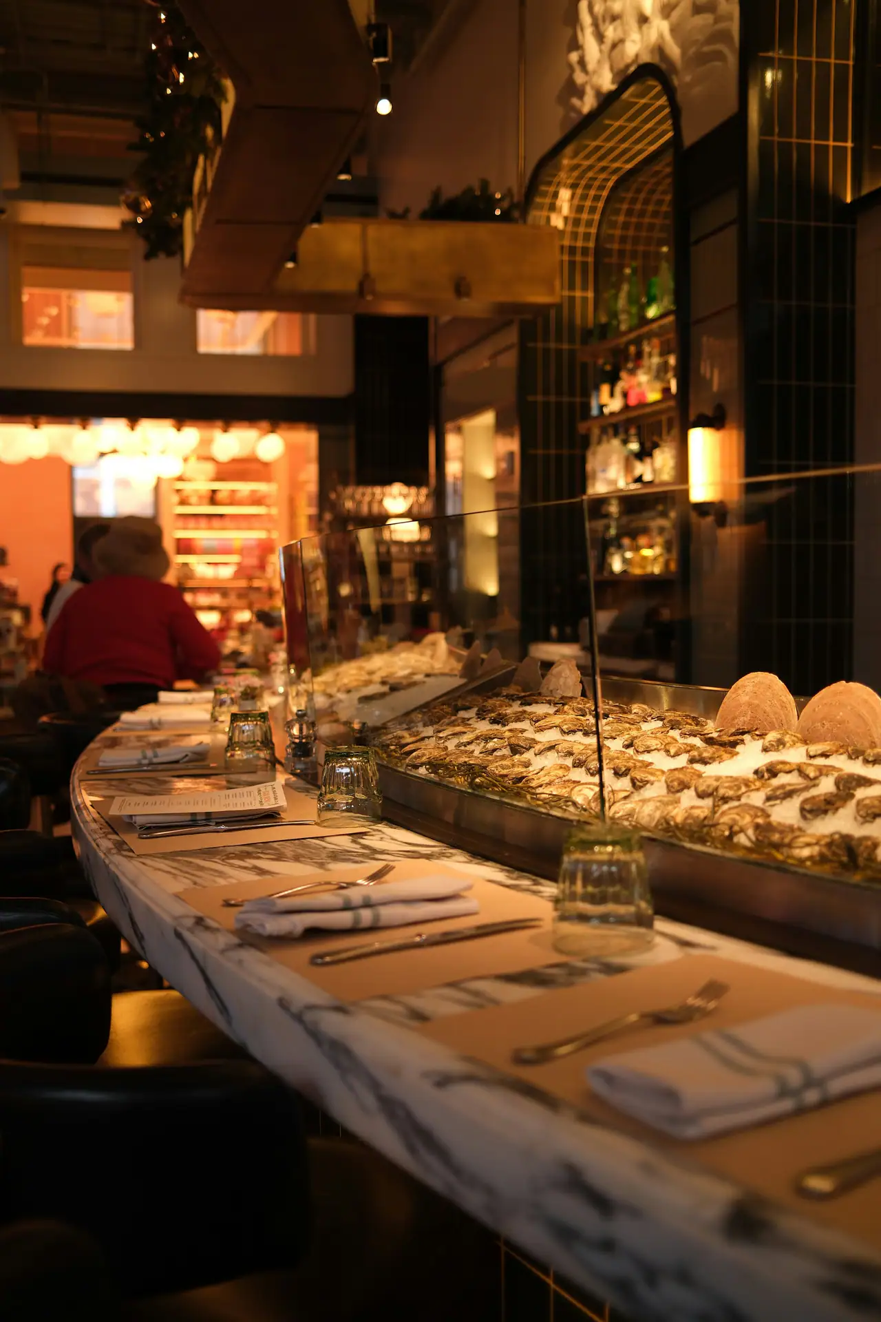 A warmly lit restaurant counter with a veined marble top, folded linen napkins and cutlery, and a tray of oysters on ice behind glass