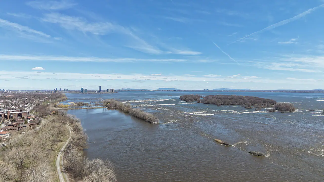 Aerial view of the St. Lawrence River at Parc des Rapides in LaSalle, with whitewater over the Lachine Rapids, wooded islets, a waterfront path along the bare-treed shore and the Montréal skyline in the distance