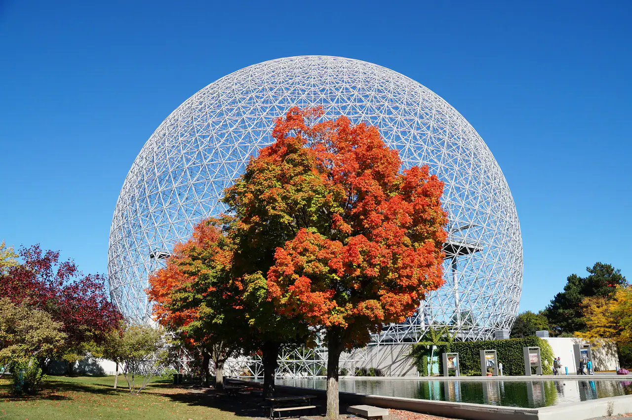 The Biosphère geodesic dome on Île Sainte-Hélène behind a maple tree in orange and red autumn leaves, with a reflecting pool in the foreground