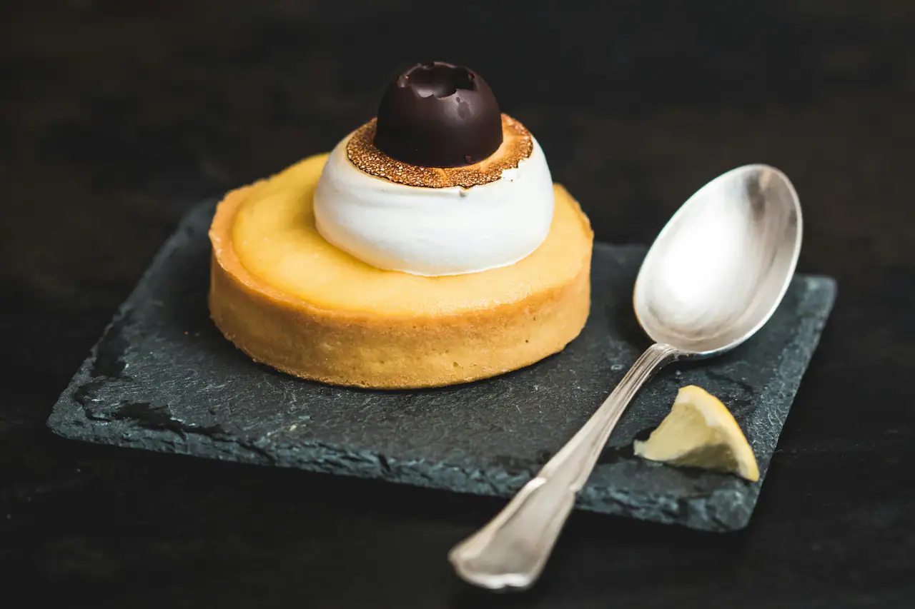 An individual lemon tart topped with toasted meringue and a dark chocolate sphere, on a slate square with a silver spoon and a lemon wedge against a dark background