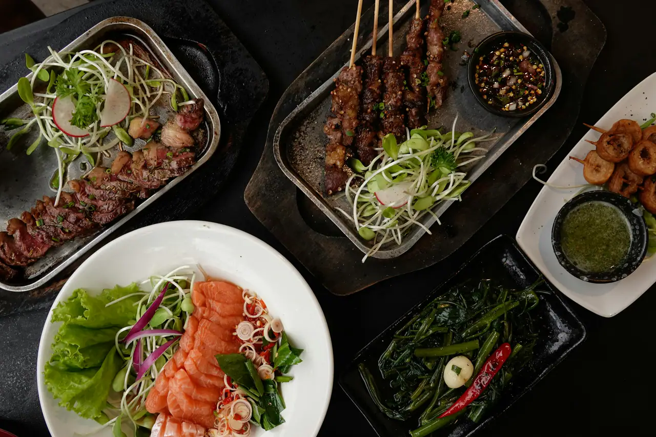 Overhead view of a dark table set with grilled skewers, sliced grilled meat, a raw fish salad with herbs and morning glory greens in trays and bowls