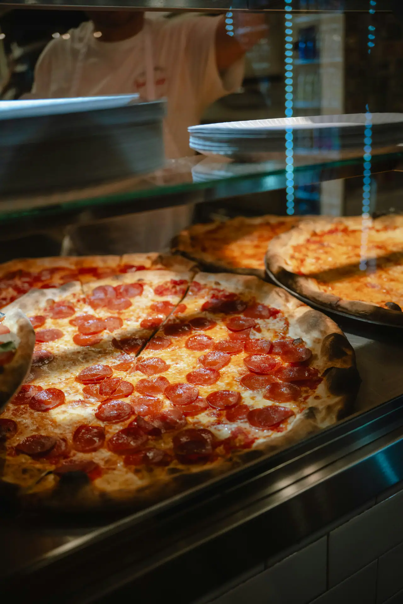 Whole pepperoni pizzas with blistered crusts displayed on a steel counter behind glass, a pizza maker blurred in the background