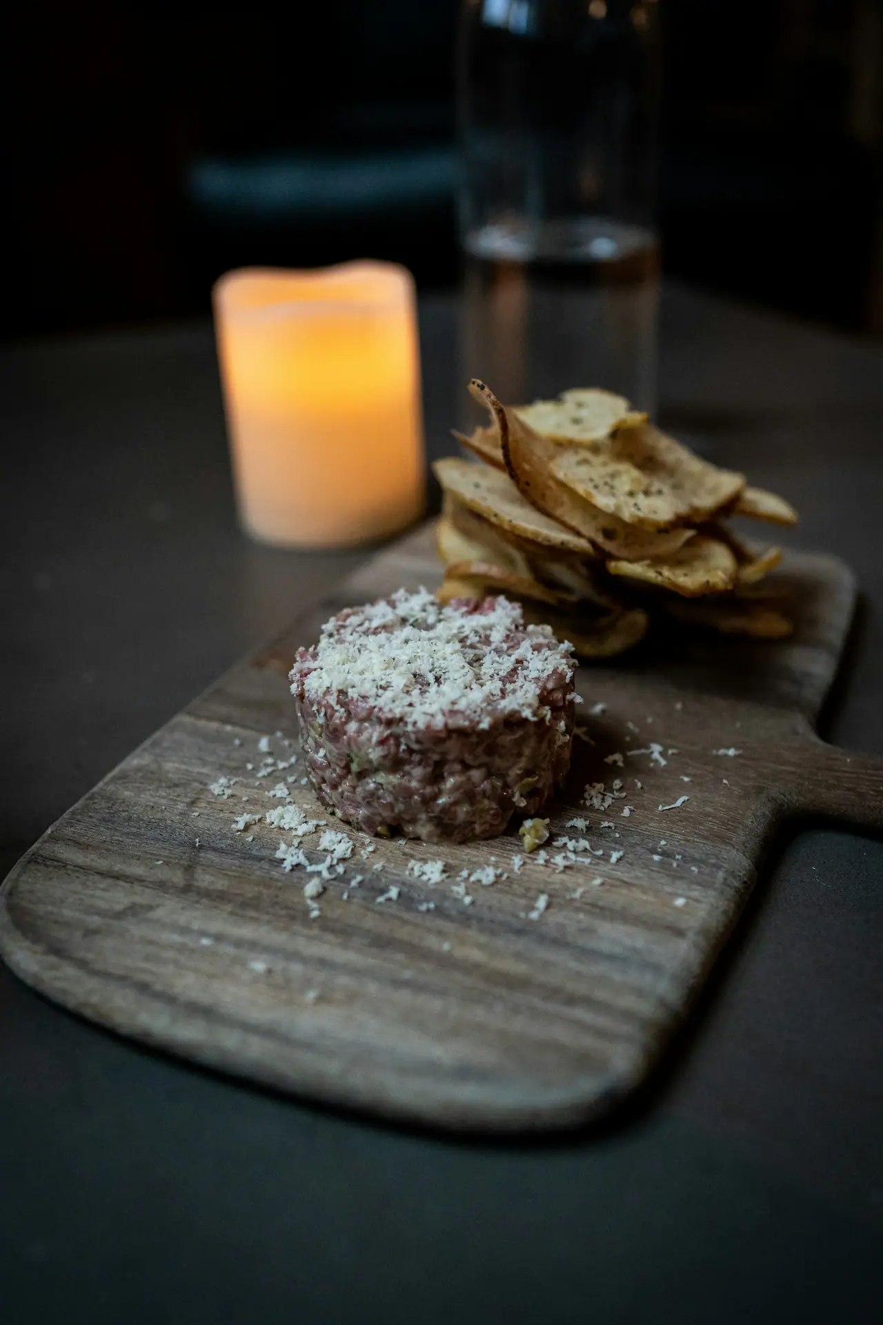 Beef tartare topped with grated cheese beside a stack of crisp potato chips on a wooden board, a candle glowing in a dark dining room behind
