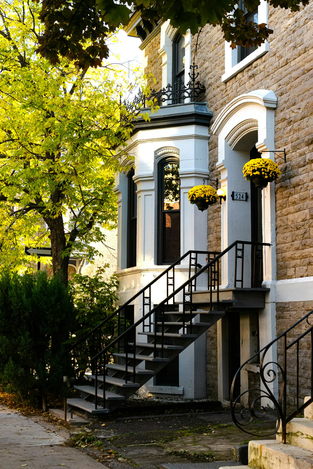 An outdoor wrought-iron staircase rising to the door of a stone-fronted Montréal-style townhouse, with hanging yellow chrysanthemums and sunlit autumn leaves beside it
