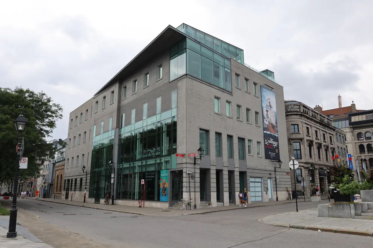 Corner of the Pointe-à-Callière museum's glass-and-stone Old Montréal building under an overcast sky, with a banner on its facade