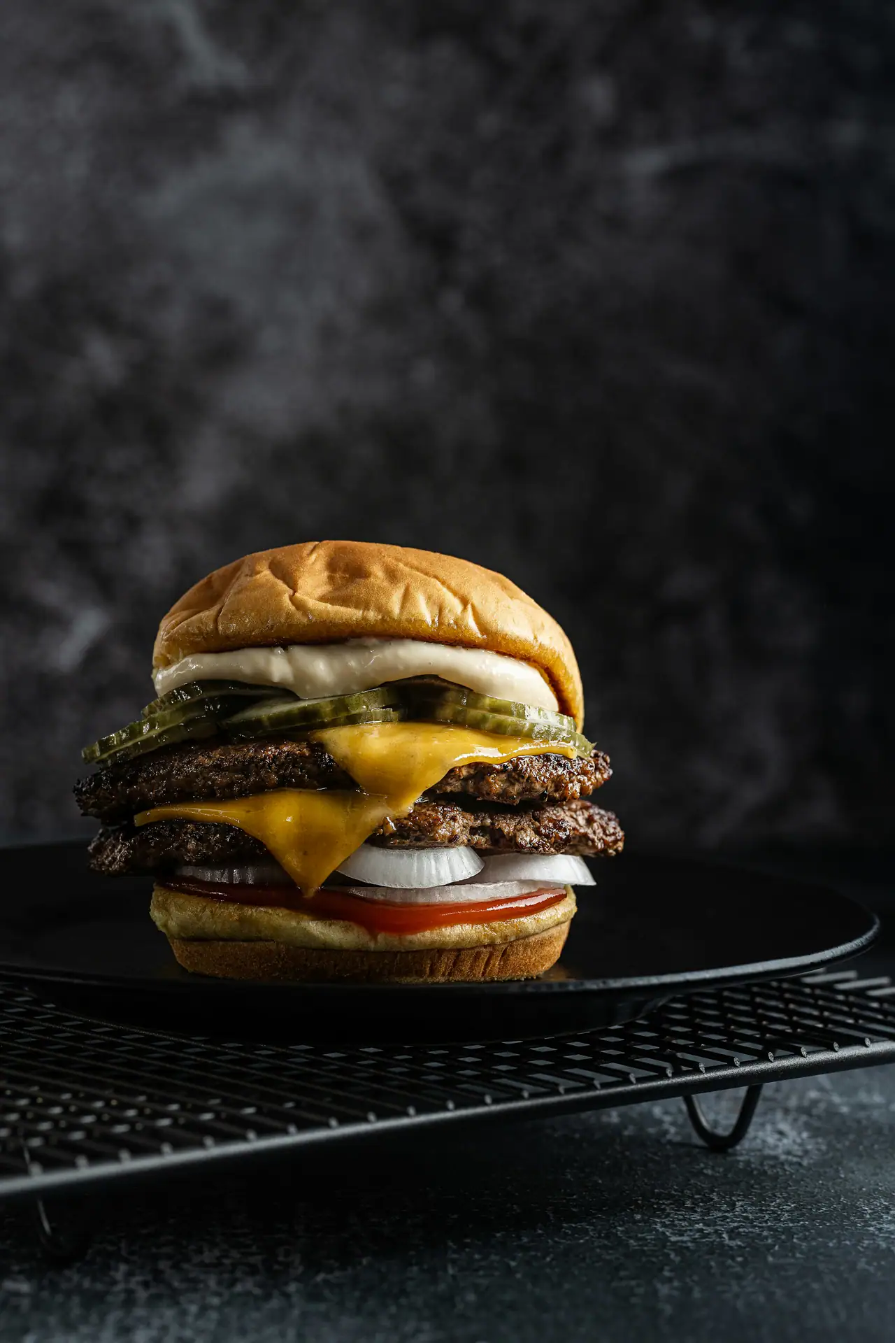 A double cheeseburger with pickles, onion and mayo on a black plate set on a wire rack against a dark, textured backdrop