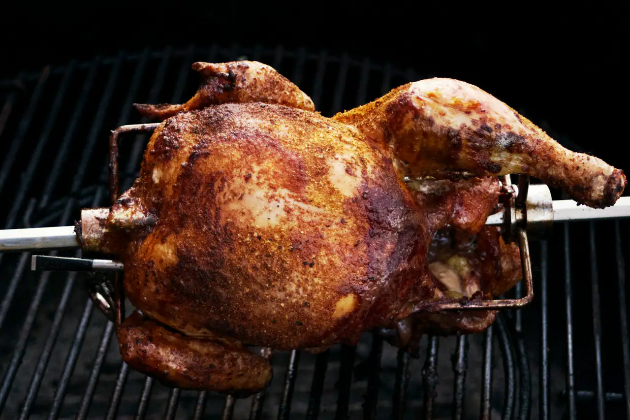 A whole spice-rubbed chicken turning on a rotisserie spit over a dark grill