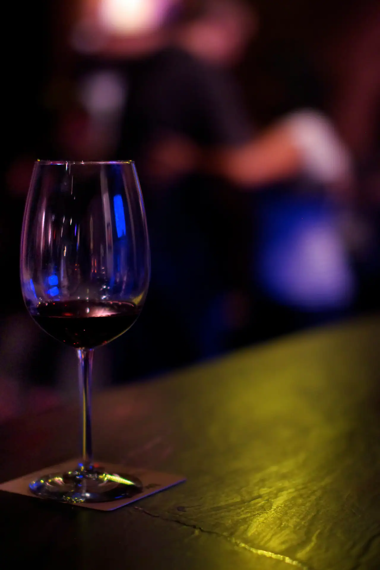 A glass of red wine on a dark bar counter, with blurred figures and blue and magenta light in the background