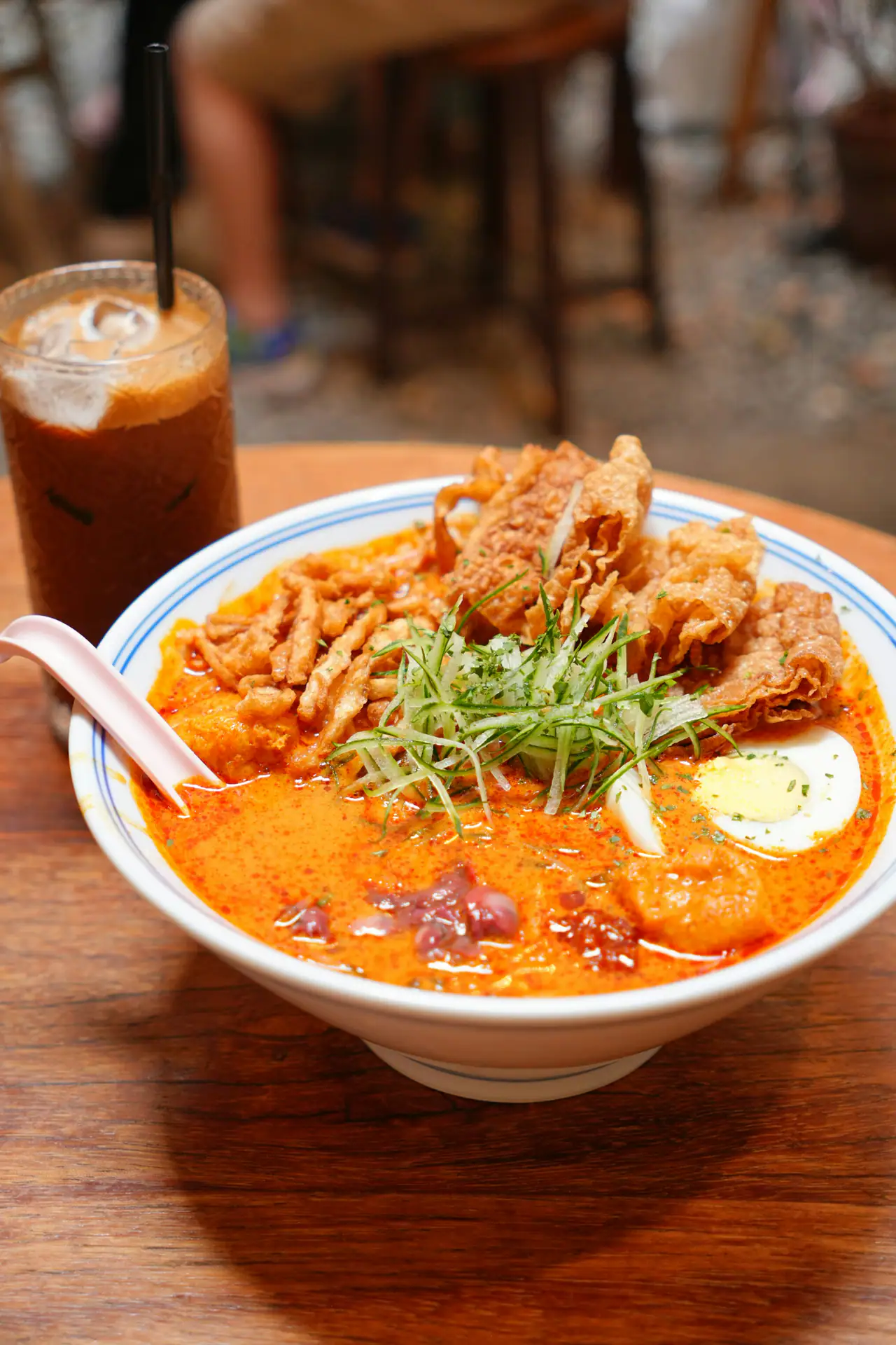 A white bowl of orange laksa noodle soup topped with crispy fried tofu, scallion shreds and a halved egg, beside an iced coffee on a wooden table