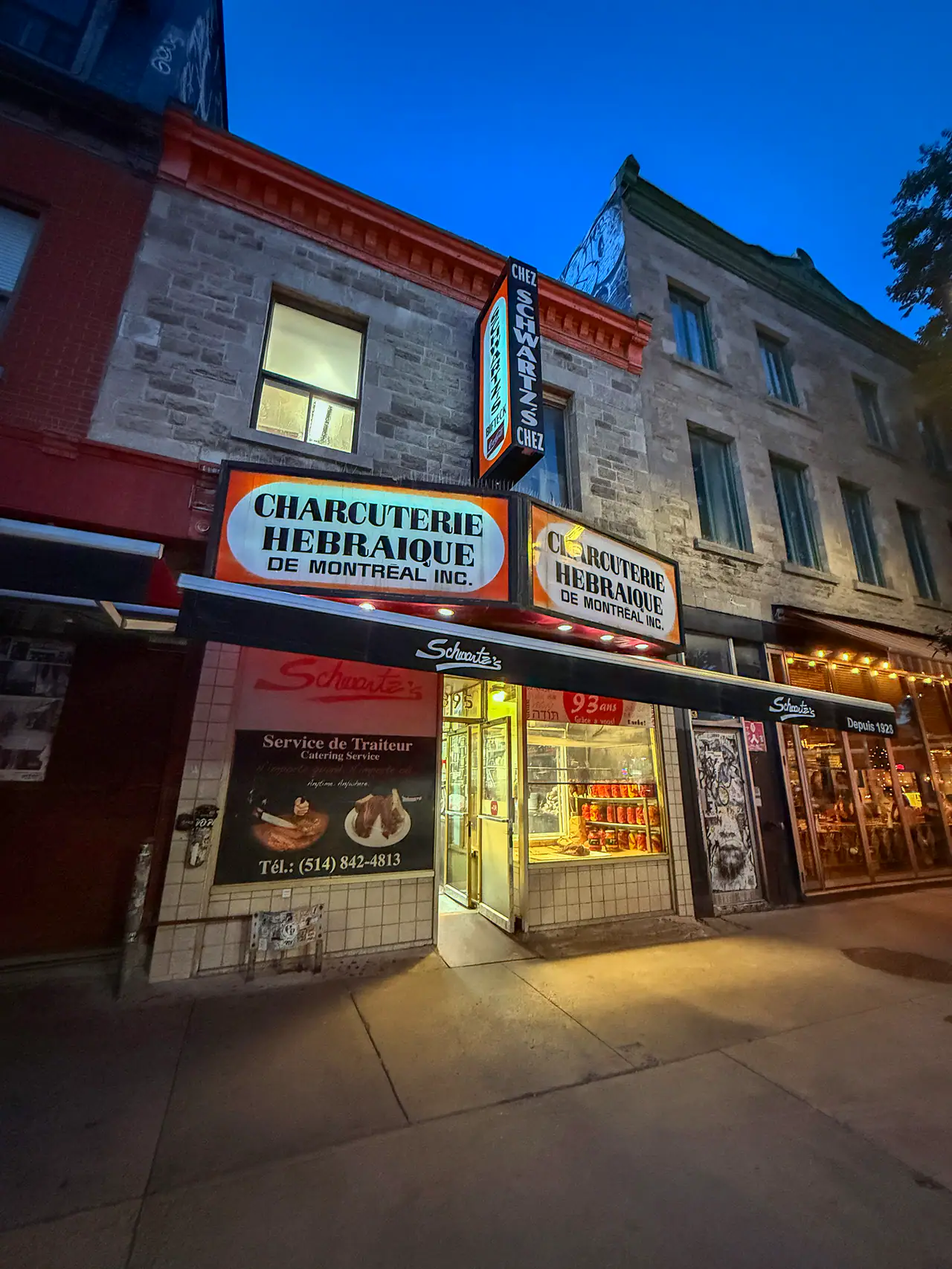 Schwartz's Deli storefront at dusk on Boulevard Saint-Laurent, its orange Charcuterie Hébraïque signs lit above the open door