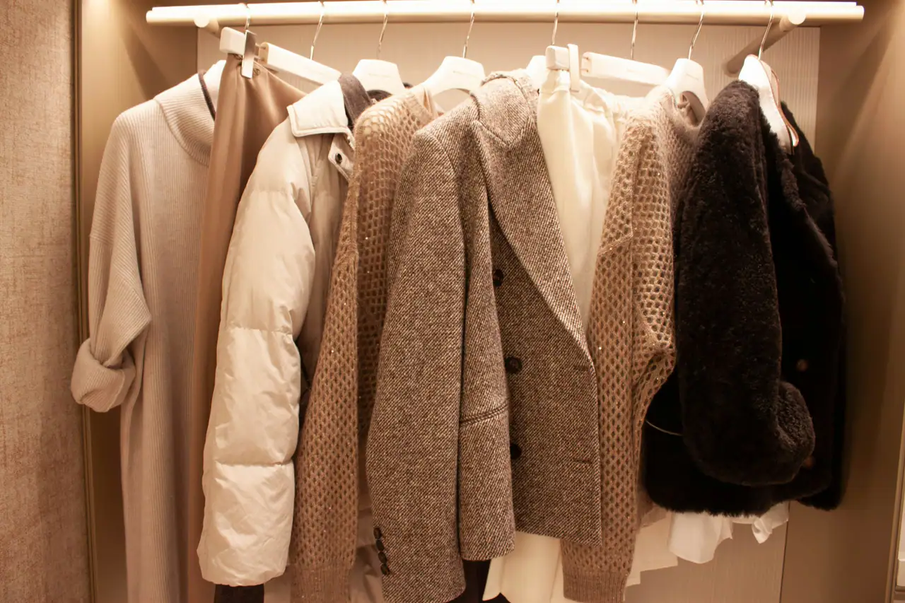 Wool, knit and shearling coats hanging on a rail in warm light, in cream, taupe and charcoal tones