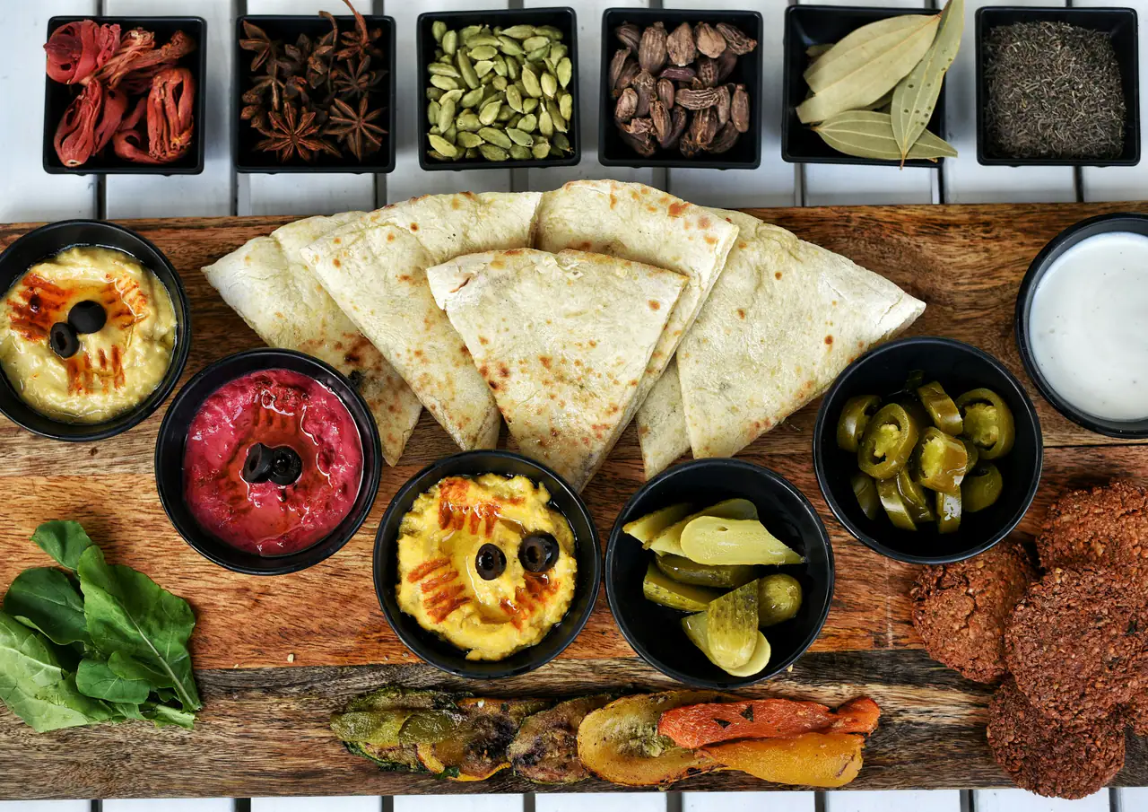 A wooden board of Middle Eastern mezze: three bowls of hummus, pickles, falafel, grilled flatbread and small dishes of whole spices, seen from above