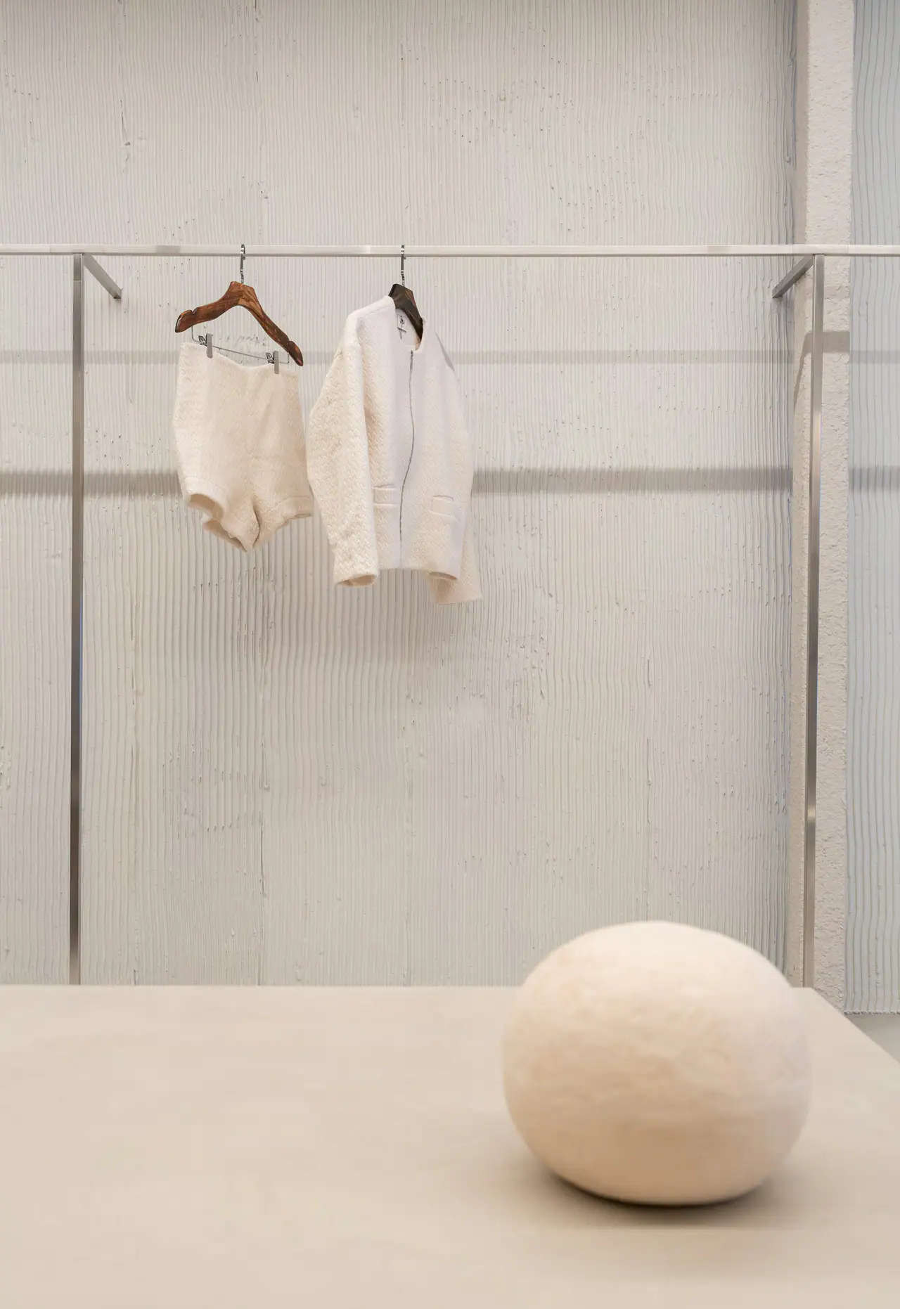 Two cream garments on hangers from a steel rail against a pale textured concrete wall, with a rounded stone-coloured sculpture on a table in the foreground