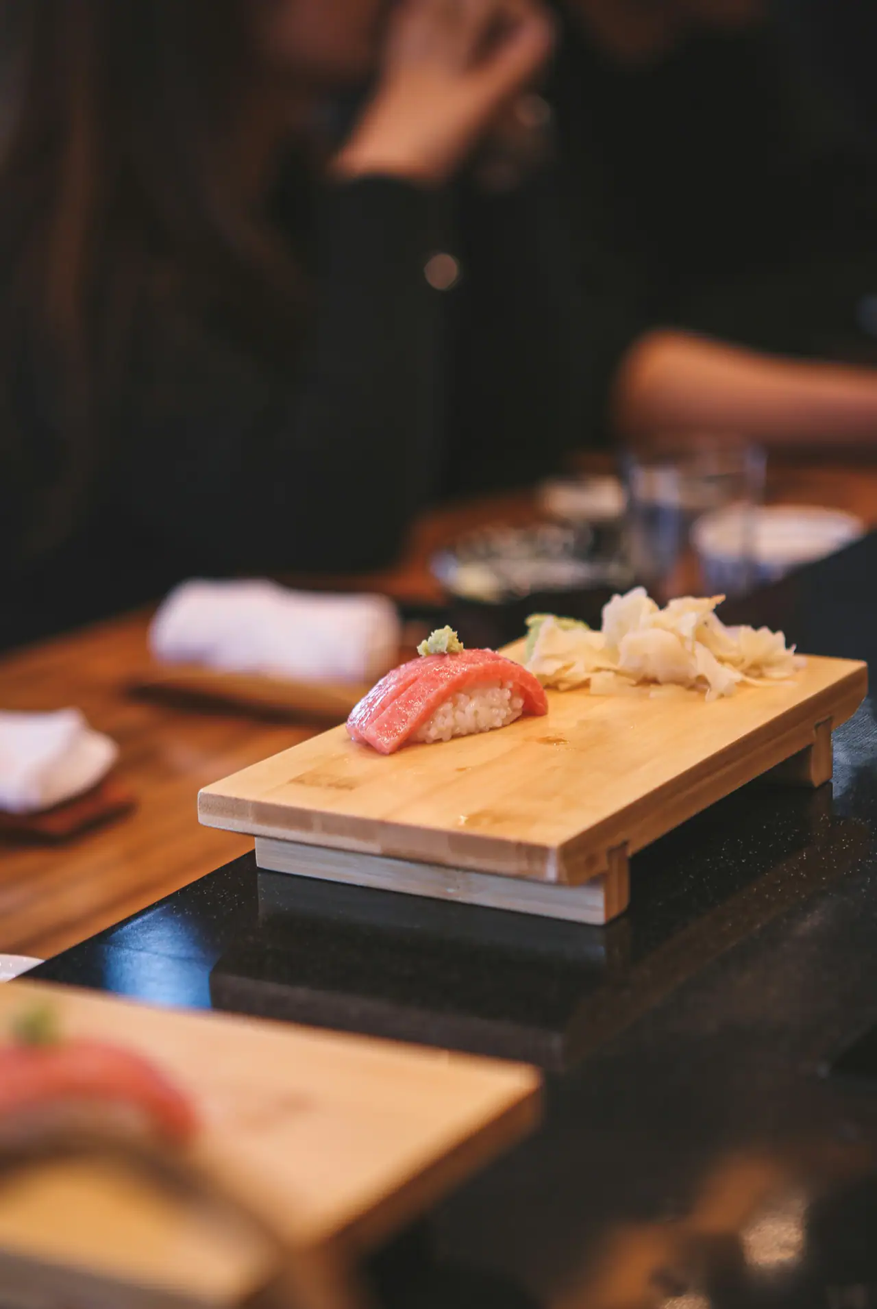A single piece of tuna nigiri with a dab of wasabi on a pale wooden board at a dark sushi counter, with pickled ginger beside it and blurred diners behind