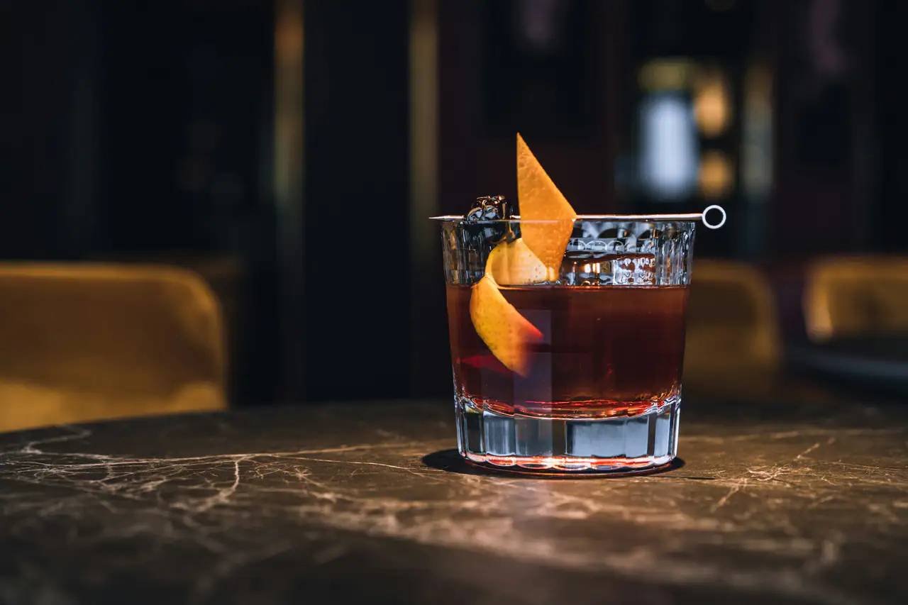 A stirred amber cocktail in a cut-crystal rocks glass with orange peel twists, on a dark veined stone bar top in a dim room