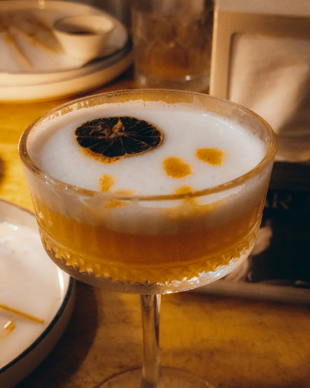 A frothy sour cocktail in a cut-crystal coupe, topped with a dried citrus slice and drops of bitters, on a warmly lit wooden table