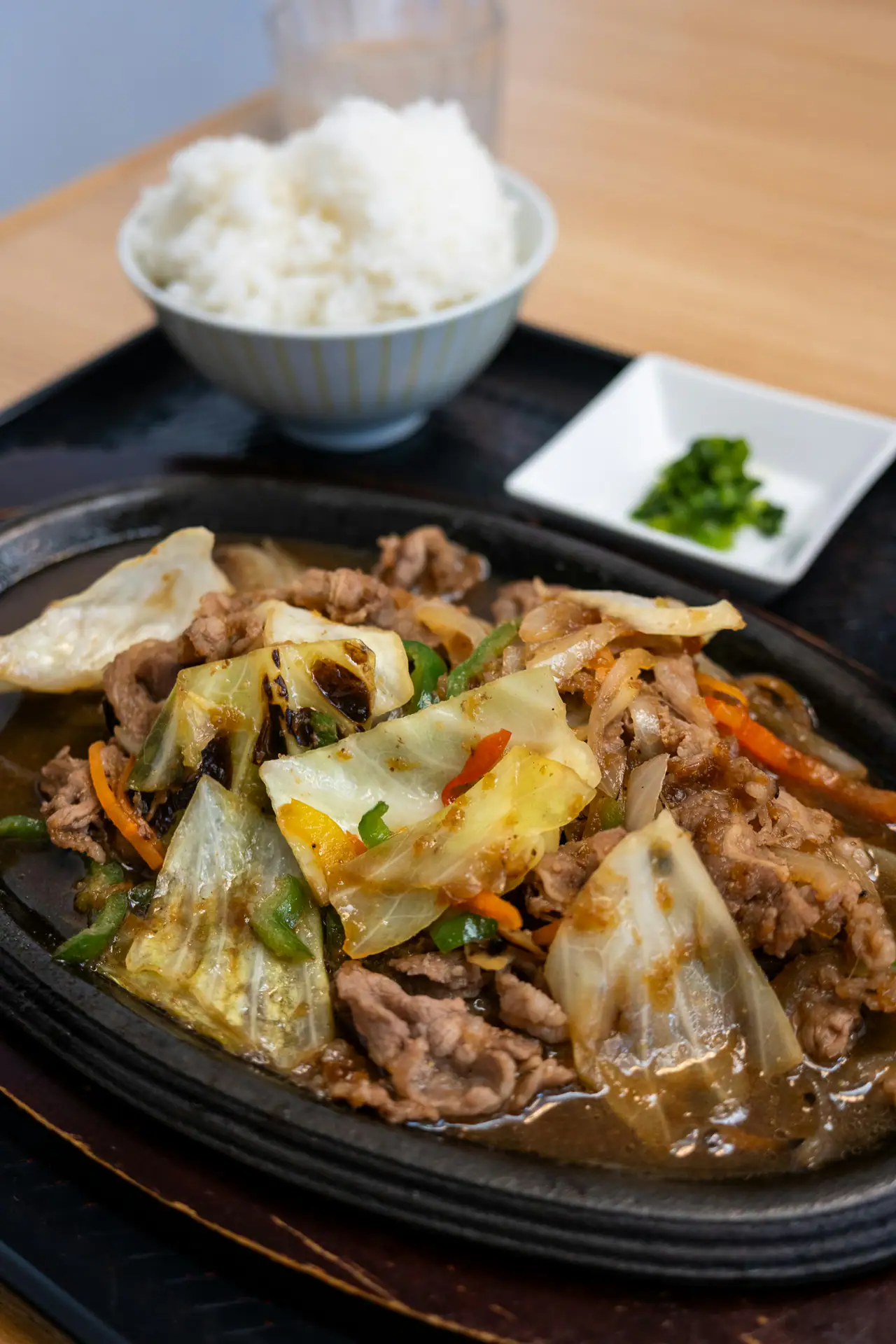 Sliced beef stir-fried with cabbage, peppers and onion on a sizzling cast-iron plate, with a bowl of white rice and chopped greens alongside